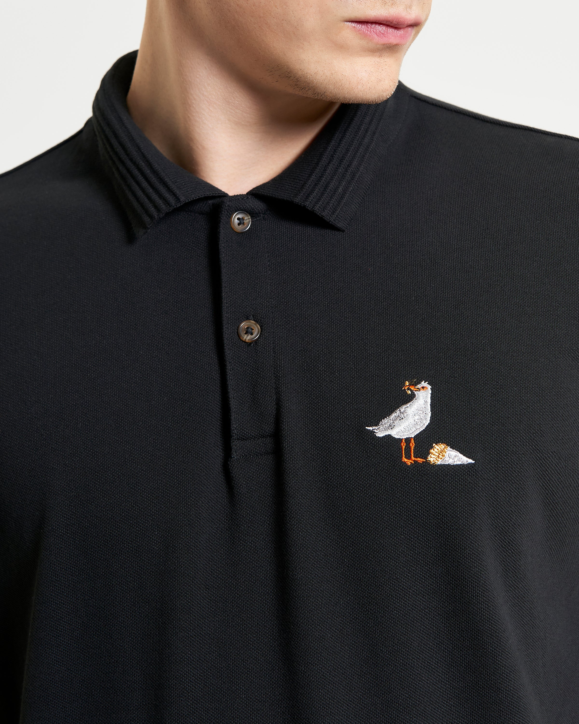 Gull Embroidery Polo – Close-up of gull embroidery on a black pique polo shirt with ribbed collar.