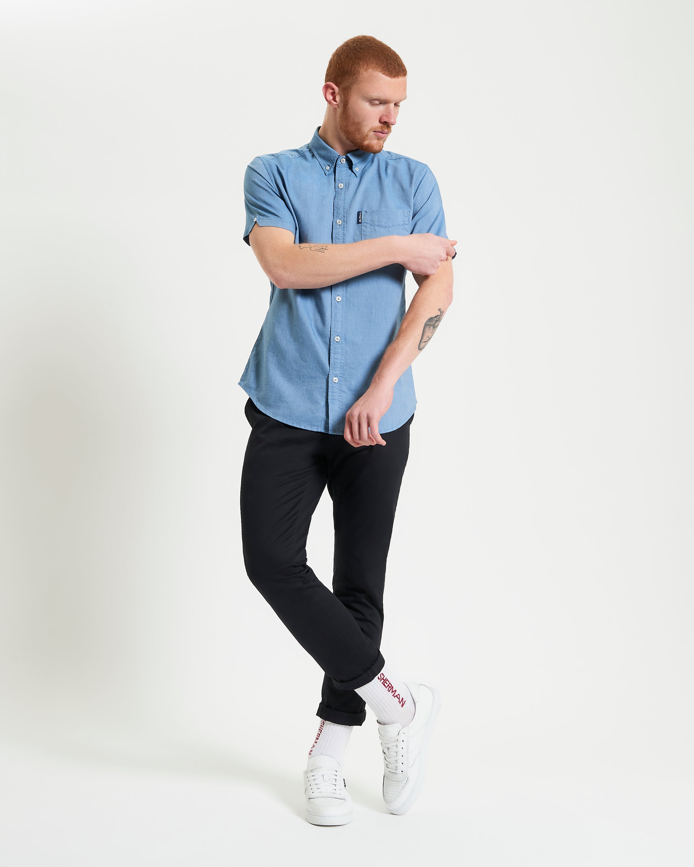 Model wearing a blue Signature Oxford Short Sleeve Shirt with black pants and white sneakers, showcasing a modern style.