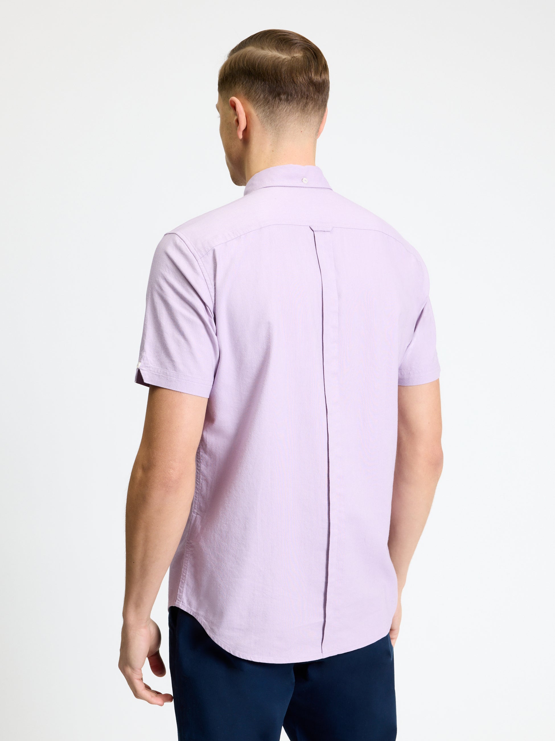 Back view of a man wearing the Signature Oxford Short Sleeve Shirt in light purple, featuring a button-down collar and back pleat.